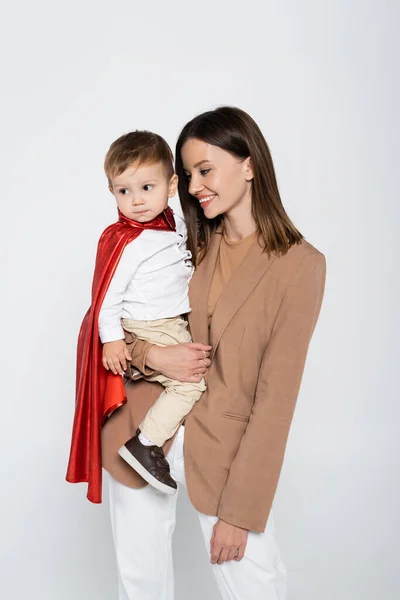 Happy mother holding in arms toddler son in superhero costume isolated on grey — Foto stock