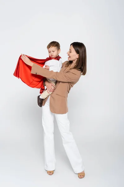Excited mother holding in arms toddler son in superhero costume on grey — стоковое фото
