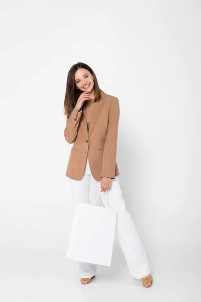 Full length of happy young woman in beige blazer holding shopping bag on white — Stock Photo