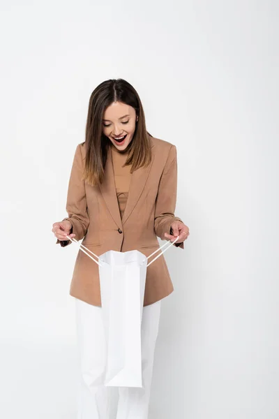 Excited young woman in beige blazer holding shopping bag on grey — Stockfoto