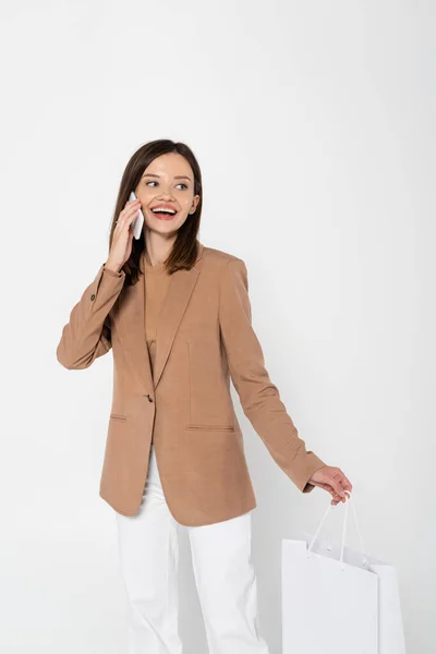 Happy young woman in beige blazer holding shopping bag and taking on smartphone on grey — Fotografia de Stock