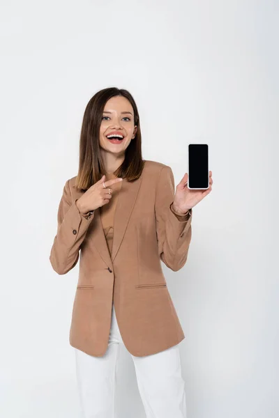 Happy young woman in beige blazer pointing at smartphone with blank screen on grey — Stock Photo