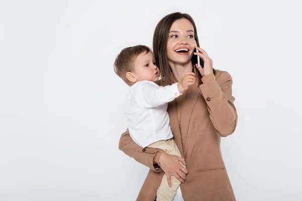 Mujer joven feliz en blazer beige hablando en el teléfono inteligente y sosteniendo en brazos niño pequeño aislado en gris - foto de stock