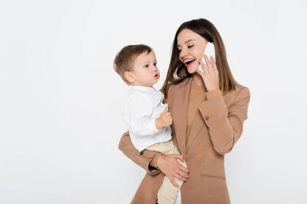 Mujer joven feliz en blazer beige hablando en el teléfono inteligente y sosteniendo en brazos niño pequeño aislado en gris - foto de stock