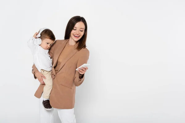 Щаслива жінка в бежевому блістері тримає смартфон і малюка хлопчика в навушниках ізольовані на сірому — стокове фото