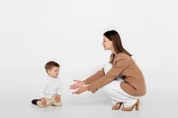 Happy woman in beige blazer sitting and reaching toddler boy on grey — Stockfoto