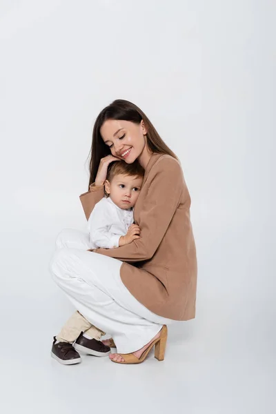 Happy woman in beige blazer sitting and hugging toddler boy on grey — Stockfoto