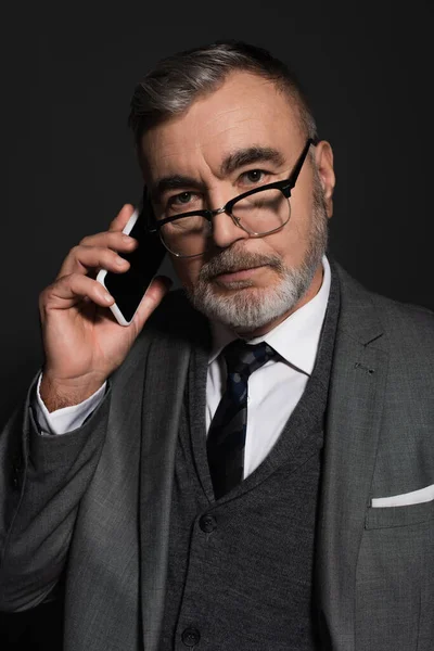 Elderly man in suit and eyeglasses looking at camera while talking on cellphone isolated on dark grey — Stock Photo