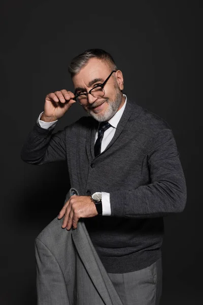 Positive senior man holding blazer and eyeglasses while smiling at camera on grey — Stockfoto