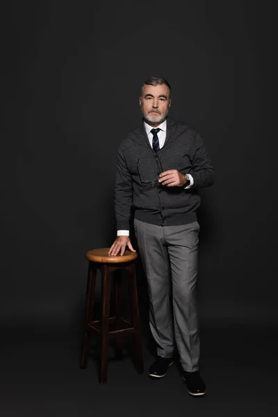 Full length view of senior businessman in stylish clothes standing near high stool on dark grey — стоковое фото