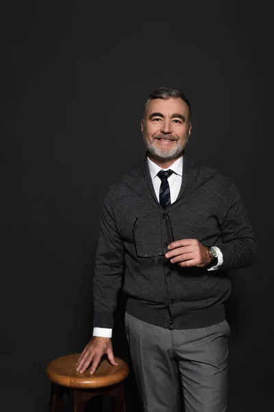 Senior businessman holding eyeglasses and looking at camera while standing near stool on dark grey — Photo de stock