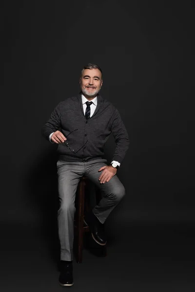 Full length view of senior man in stylish clothes sitting on high stool and smiling at camera on grey — Photo de stock