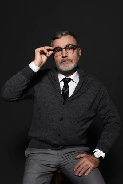 Bearded senior man looking at camera while adjusting eyeglasses on dark grey — стоковое фото