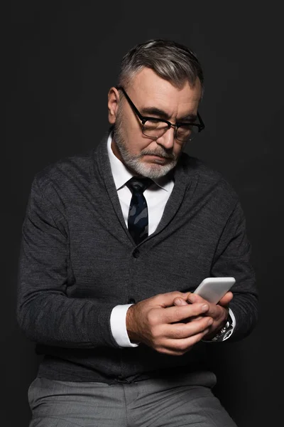 Senior man in jumper and eyeglasses using mobile phone isolated on black — Stock Photo