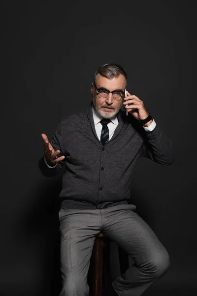 Серйозний старший чоловік вказує рукою і розмовляє по мобільному телефону, сидячи на темно-сірому — стокове фото