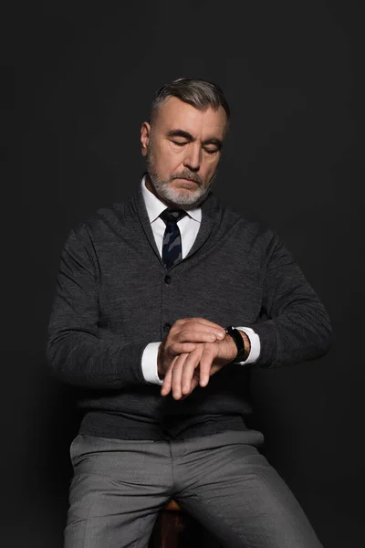 Senior man in jumper and tie looking at wristwatch on dark grey — Photo de stock