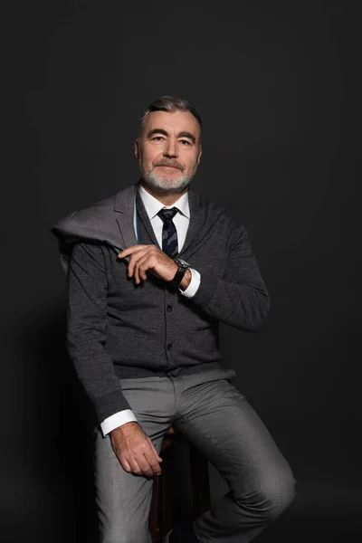 Positive senior businessman holding blazer while sitting and looking at camera on grey — Fotografia de Stock