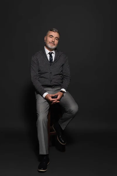 Full length view of smiling grey-haired man in stylish clothes sitting on high stool on dark grey — Foto stock