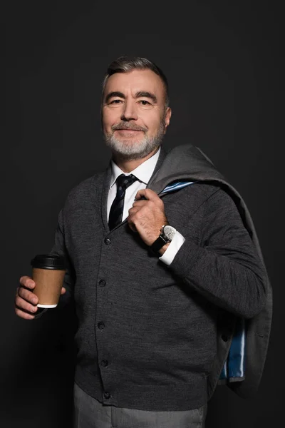 Bearded man holding coffee to go and blazer while smiling at camera on grey — Stockfoto