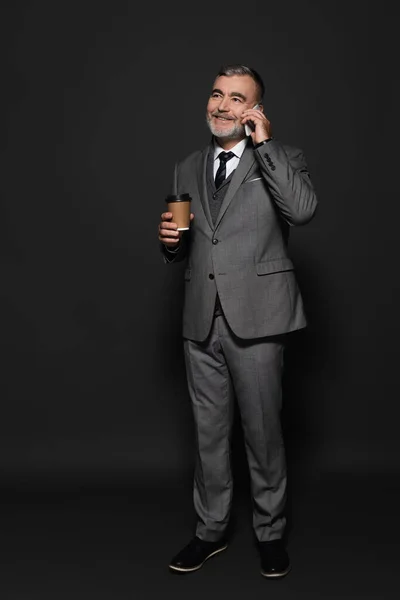 Full length view of smiling senior businessman with takeaway drink talking on mobile phone on dark grey — Foto stock