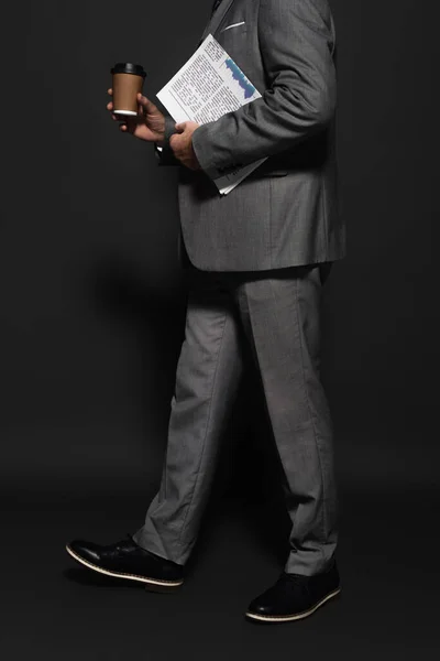 Partial view of businessman in suit walking with newspaper and takeaway drink on dark grey — Stock Photo