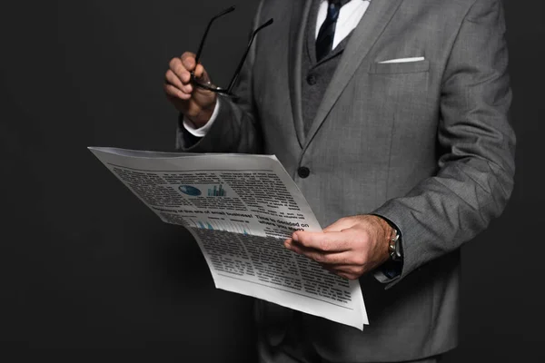 Cropped view of businessman holding eyeglasses and reading newspaper on dark grey - foto de stock