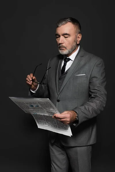 Senior businessman in suit holding eyeglasses while reading newspaper on dark grey — Foto stock