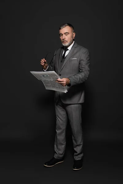 Full length view of elderly man in formal wear holding eyeglasses while reading newspaper on dark grey — Stock Photo