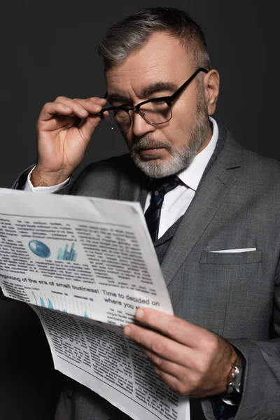 Senior man in formal wear adjusting eyeglasses while reading newspaper isolated on dark grey — Foto stock