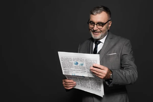 Happy senior man in suit and eyeglasses reading newspaper isolated on dark grey — Foto stock