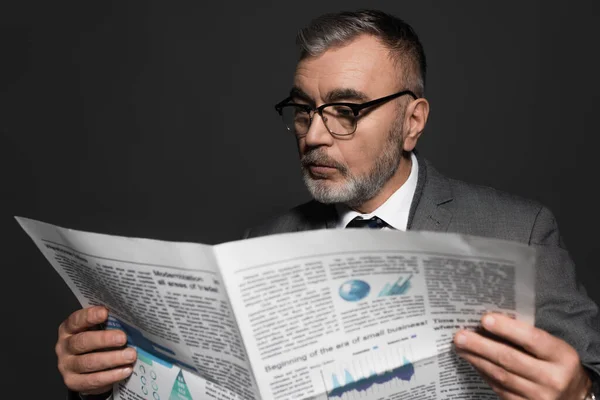 Bearded senior businessman in eyeglasses reading blurred newspaper isolated on dark grey — Fotografia de Stock