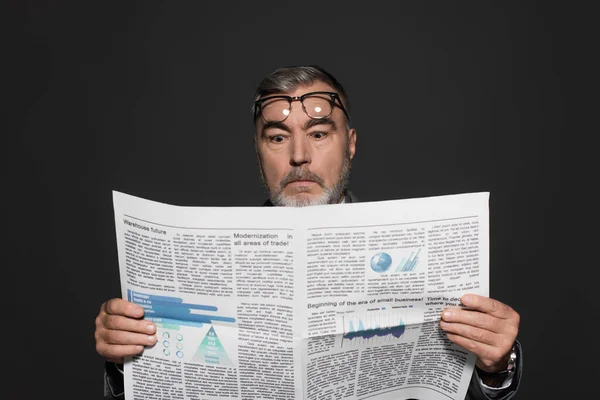 Surprised senior businessman with eyeglasses on forehead reading newspaper isolated on dark grey — Photo de stock