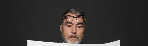 Shocked man with eyeglasses on forehead reading news isolated on dark grey, banner — Fotografia de Stock