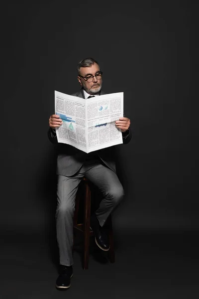 Full length view of man in suit and eyeglasses sitting on high stool and reading newspaper on dark grey - foto de stock