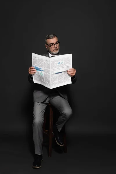 Full length view of man in suit and eyeglasses sitting on high stool and reading newspaper on dark grey — стоковое фото