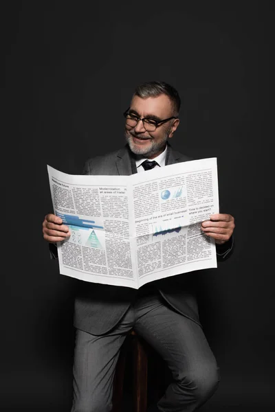 Cheerful man in suit sitting on stool and reading newspaper on dark grey — Stockfoto