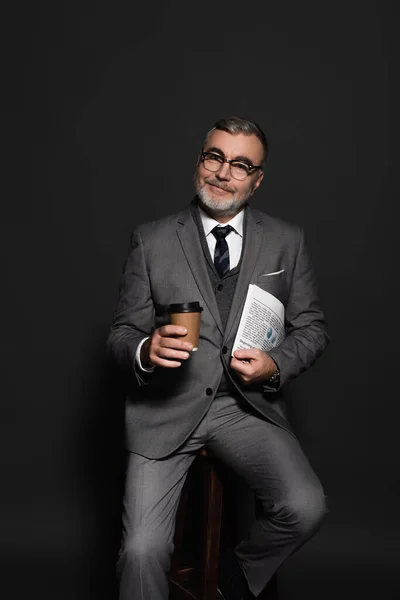 Стильний старший бізнесмен посміхається на камеру, сидячи з кавою, щоб піти і газети на темно-сірому — стокове фото