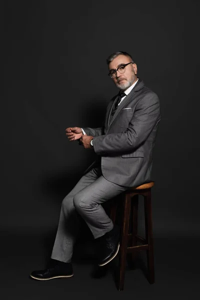 Full length view of senior man in suit and eyeglasses sitting on high stool and looking at camera on dark grey - foto de stock