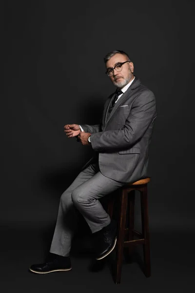 Full length view of senior man in suit and eyeglasses sitting on high stool and looking at camera on dark grey — Stock Photo