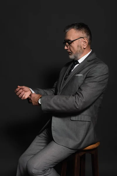 Senior man adjusting sleeve of blazer while sitting on stool on dark grey — Foto stock