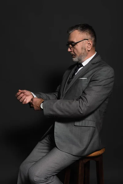 Senior man adjusting sleeve of blazer while sitting on stool on dark grey — стоковое фото