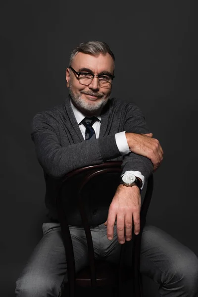 Pleased senior man in jumper and eyeglasses sitting on chair and looking at camera on dark grey - foto de stock