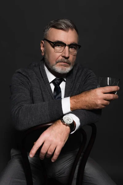 Serious and stylish senior man sitting on chair with glass of whiskey isolated on dark grey — Photo de stock