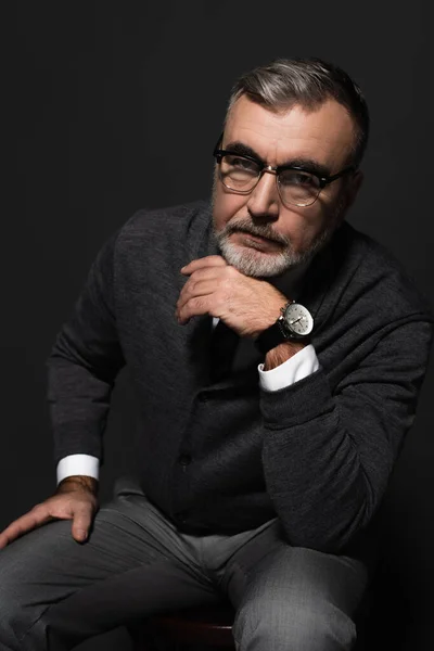 Serious senior man in jumper, eyeglasses and wristwatch looking at camera while sitting on dark grey — стоковое фото