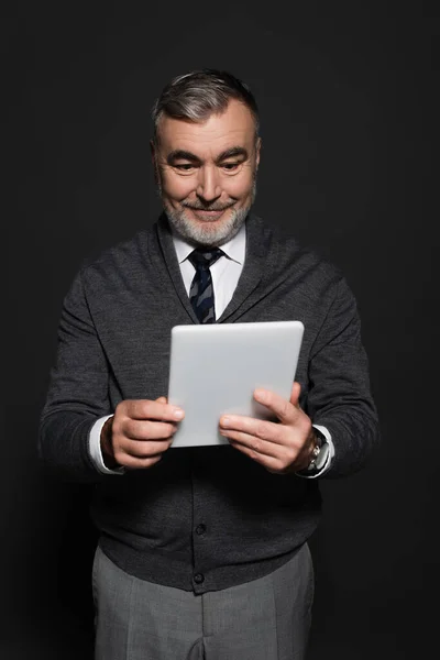 Joyful bearded man in jumper and tie looking at digital tablet on dark grey — стоковое фото