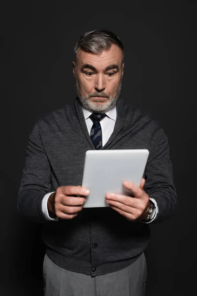 Surprised senior man in jumper and tie looking at digital tablet isolated on dark grey — Stockfoto