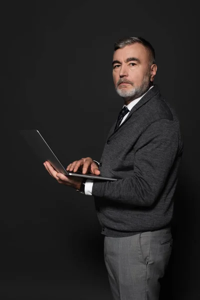 Senior bearded man in jumper holding laptop and looking at camera on dark grey - foto de stock