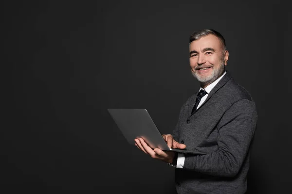 Happy senior man with laptop looking at camera isolated on dark grey — Stock Photo