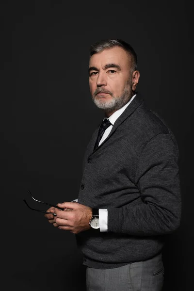 Grey-haired senior man in stylish clothes holding eyeglasses while looking at camera on dark grey — Photo de stock