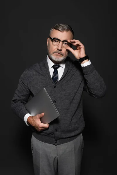 Stylish senior man with laptop adjusting eyeglasses and looking at camera on dark grey - foto de stock